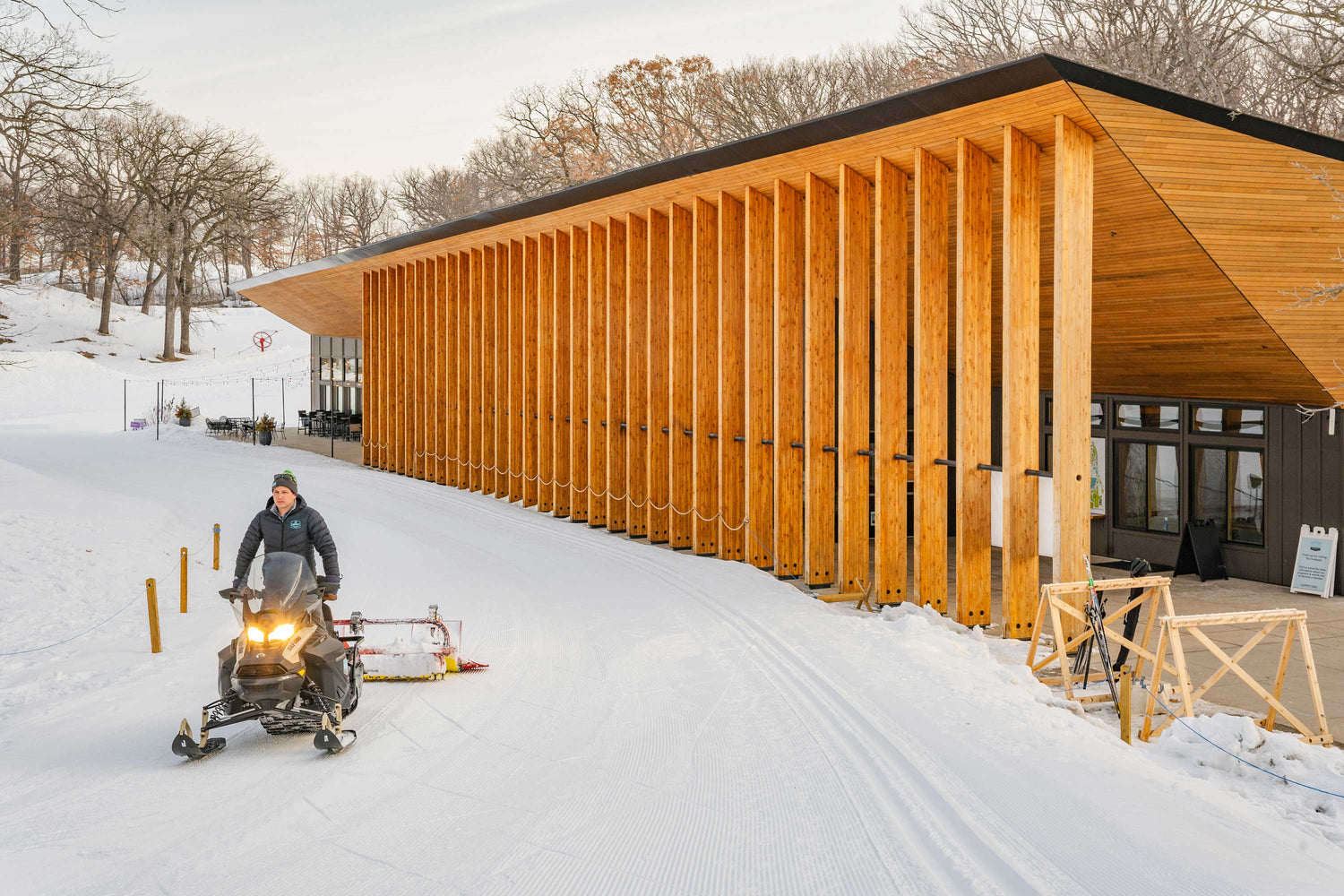 Person using a snowmobile in front of a modern wooden building in a snowy landscape.